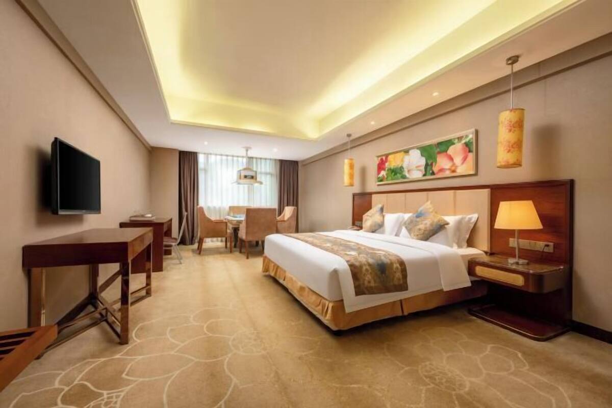 Habitación de hotel con cama, mesita de noche, lámpara, televisión, escritorio, silla y un cuadro en la pared.