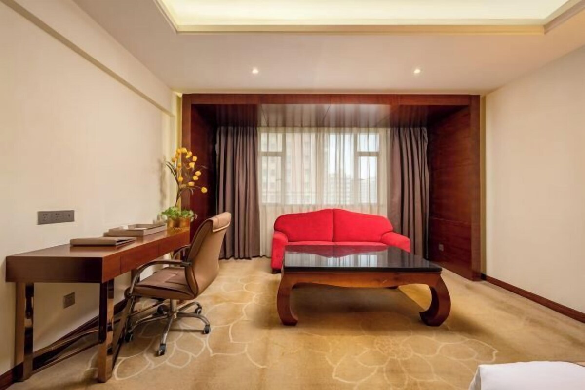 Ein Hotelzimmer mit einem Holztisch, einem roten Sofa, einem Couchtisch und einem großen Fenster mit Vorhängen.