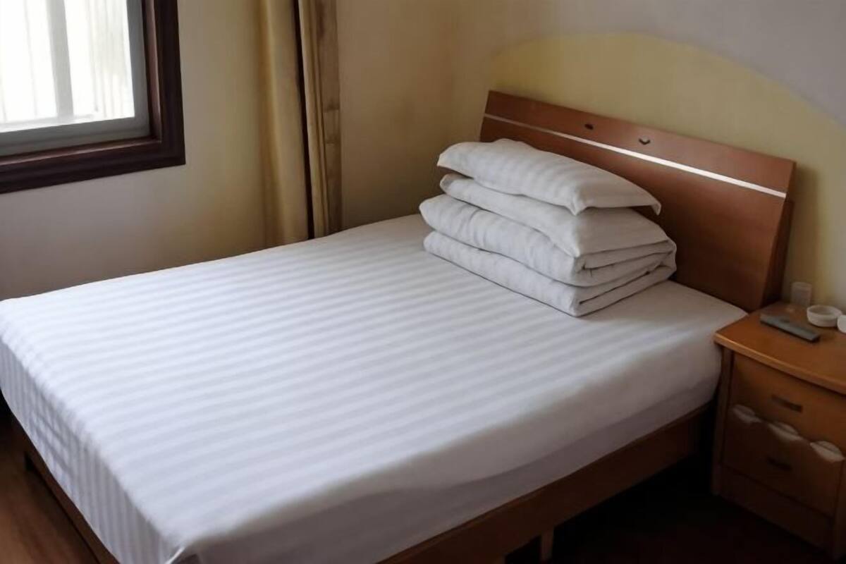 A neatly made bed with pillows, a wooden headboard, a bedside table with a lamp, and a window with curtains.