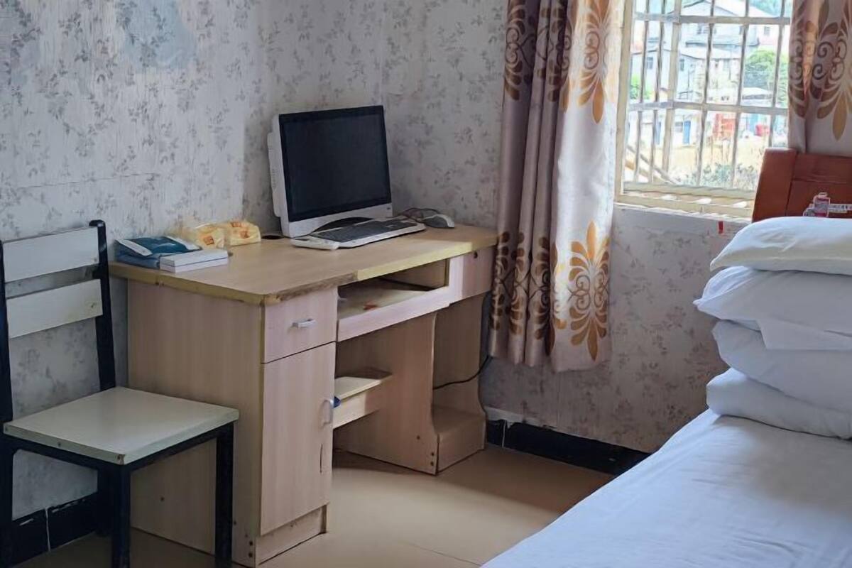A bedroom with a bed, a desk with a computer, a chair, and a window with curtains.