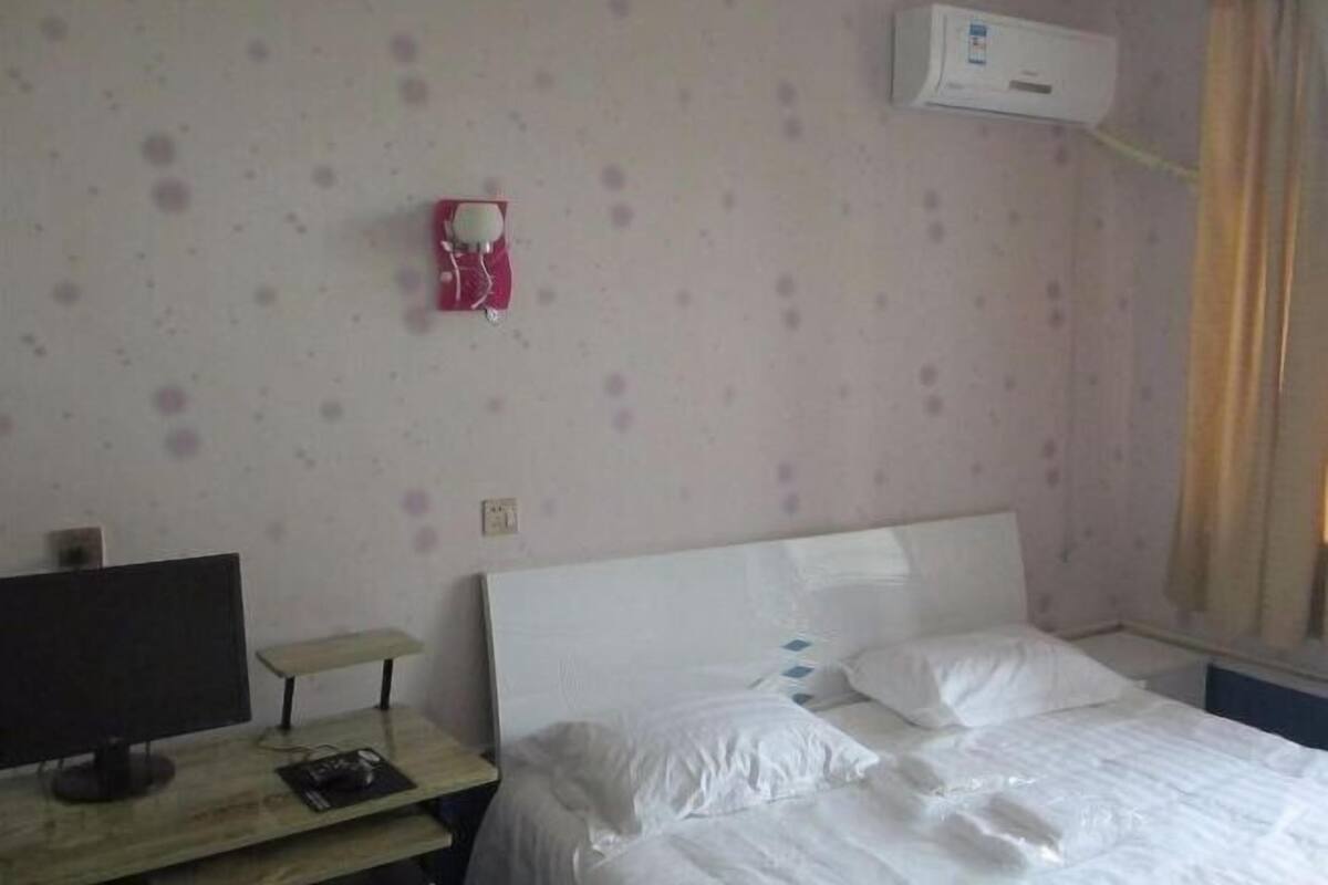 A hotel room with a bed, a desk with a computer, and a wall-mounted air conditioner.