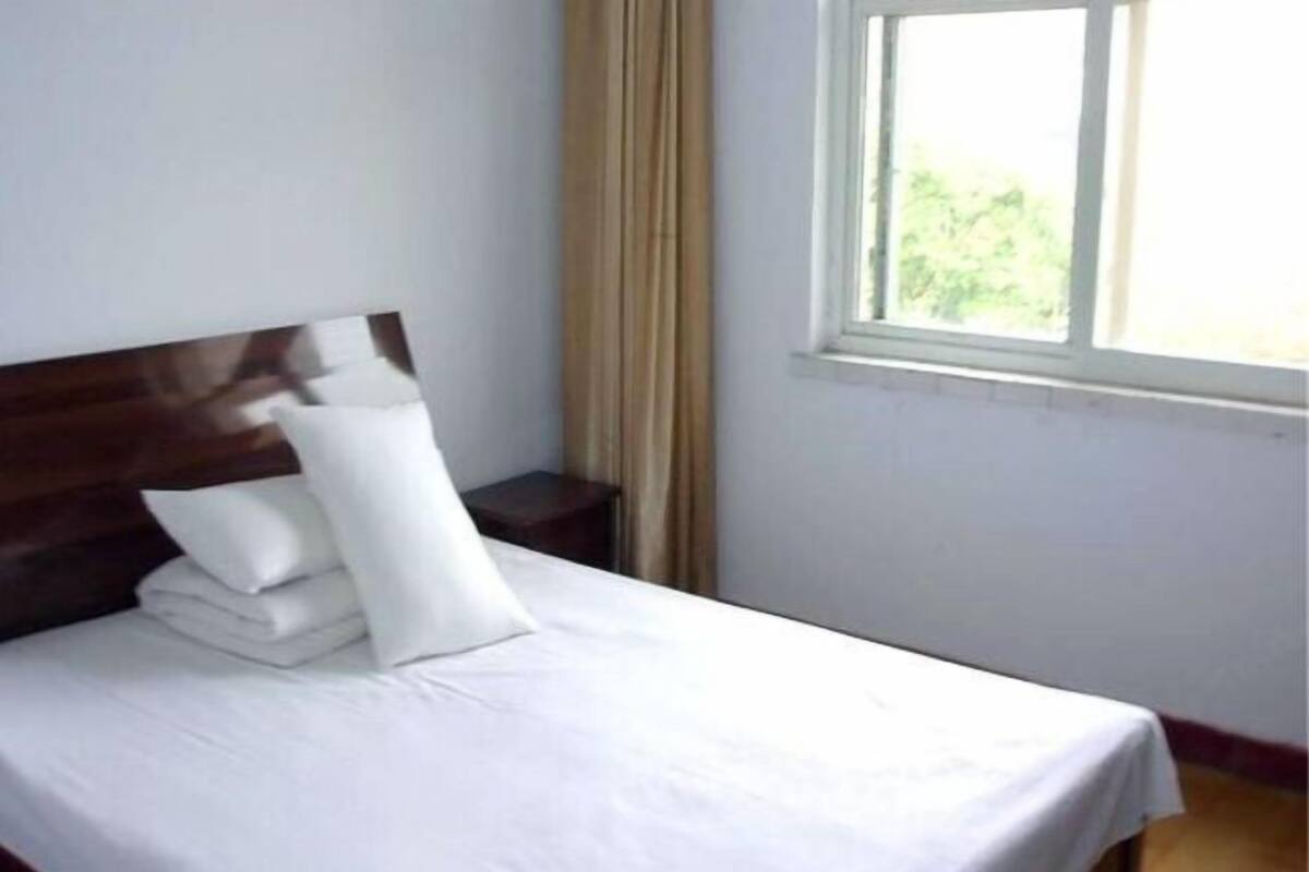 A neatly made bed with white linens and a wooden headboard in a simple room with a window and curtains.