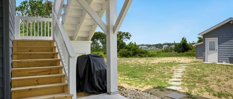 A covered outdoor staircase leading to a wooden deck, with a covered grill and a house in the background.