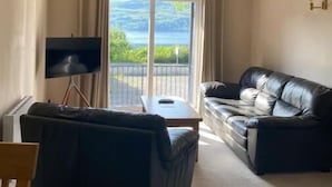 Una sala de estar con un sofá de cuero negro, una mesita y un televisor sobre un trípode. Cuenta con un balcón con vistas a la vegetación y una masa de agua.