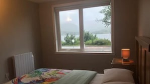Un dormitorio con cama, una ventana con vista a una masa de agua y vegetación, y una lámpara de noche.