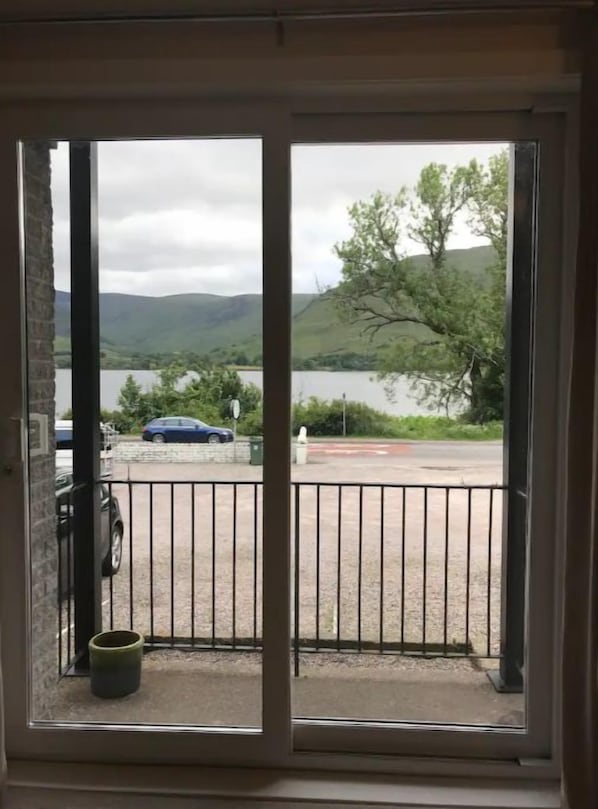 A view from a window with a metal railing, overlooking a parking area and a body of water with hills in the background. - Borrodale, Lochside Apartments (Fort William)