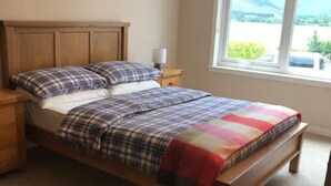 Un dormitorio con cama de madera, mesita de noche, ventana con vistas a zonas verdes y piso alfombrado.
