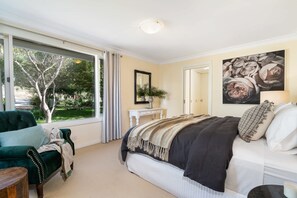 A bedroom with a large bed, a window with curtains, a bedside table with a plant, and a wall-mounted mirror.