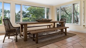 A dining area with a wooden table, benches, wicker chairs, and large windows with a view of trees. - Sandhurst - Stunning Retreat - Pool & Tennis Court (Orange)