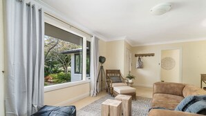 A living room with a sofa, ottoman, and a chair with a cushion. There is a window with curtains, a ceiling light, and a wall-mounted decorative item. - Sandhurst - Stunning Retreat - Pool & Tennis Court (Orange)
