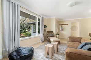 Un salon comprenant un canapé, un pouf et un fauteuil avec un coussin. On y trouve une fenêtre avec des rideaux, un luminaire au plafond et un objet décoratif fixé au mur.
