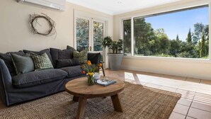 A living room with a dark grey sofa, a round wooden coffee table, and a large window with a view of trees. - Sandhurst - Stunning Retreat - Pool & Tennis Court (Orange)