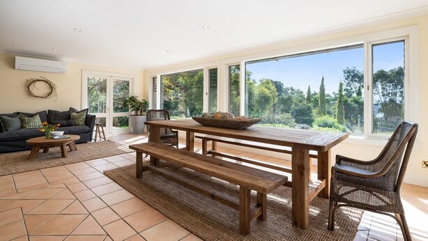 A spacious living area with a large wooden table, a sofa, and a view of a garden through large windows. - Sandhurst - Stunning Retreat - Pool & Tennis Court (Orange)