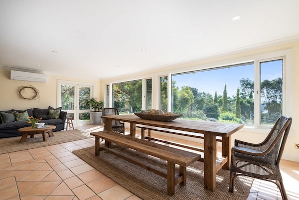 Un vaste espace de vie comprenant une grande table en bois, un canapé et une vue sur un jardin grâce à de grandes fenêtres.
