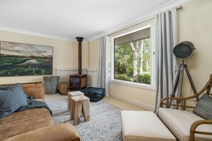 A cozy living room with a fireplace, a large window with a view of trees, a painting on the wall, and a wicker chair.