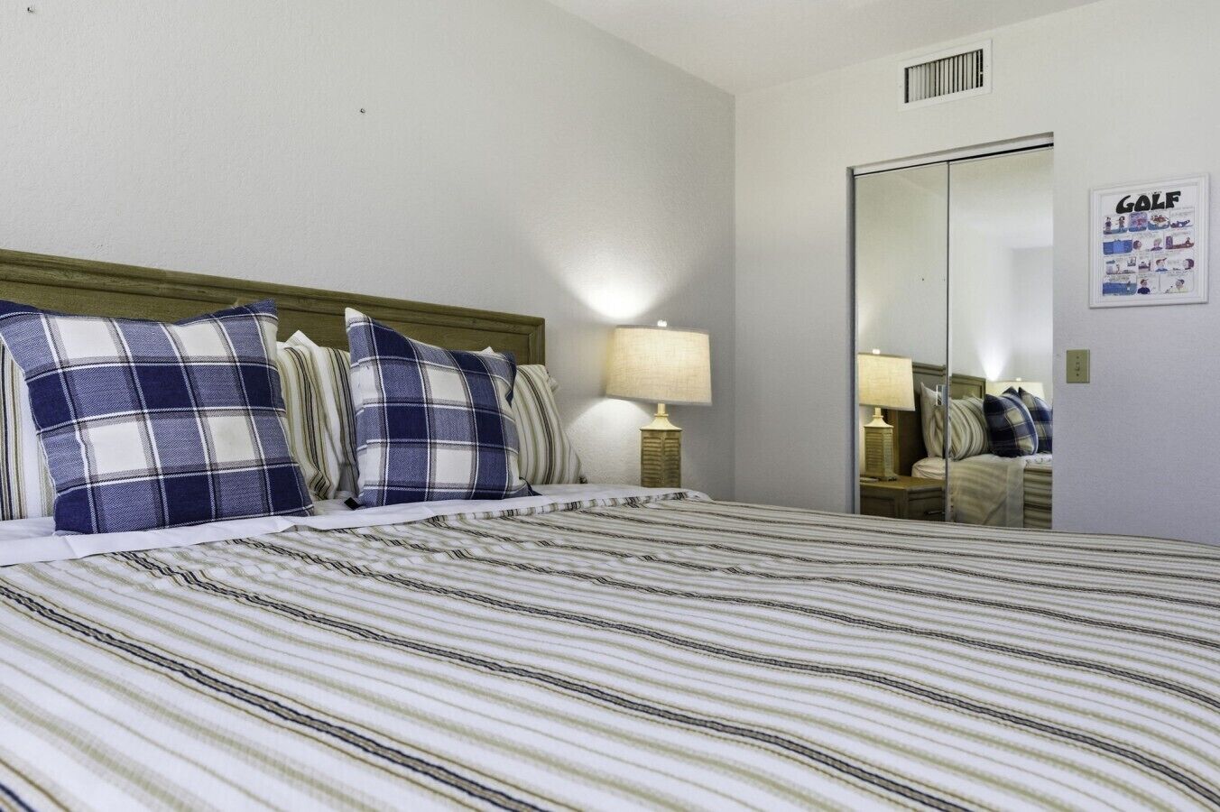 Une chambre d’hôtel avec un lit, deux oreillers, une table de chevet, une lampe, un miroir et une affiche accrochée au mur.