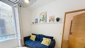 Quarto com sofá azul, uma pequena mesa branca, porta de madeira e janela com persianas.