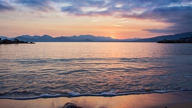 Uma praia com águas calmas e montanhas ao longe sob um céu de pôr do sol.