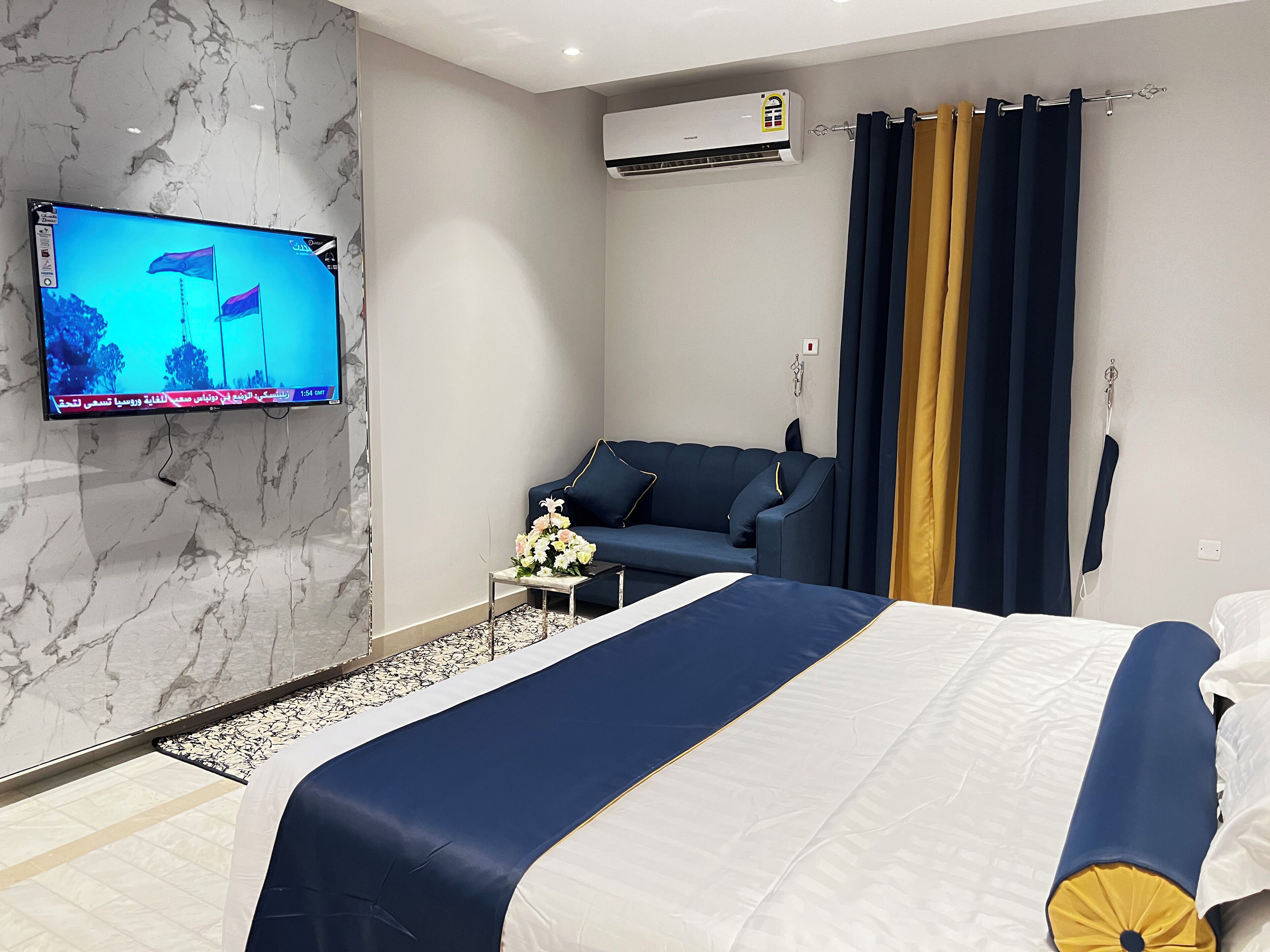 A hotel room with a bed, a sofa, a TV mounted on the wall, and an air conditioning unit.
