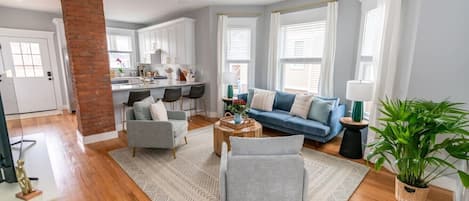 Een ruime woonkamer met een blauwe bank, twee fauteuils, een salontafel en een haard van baksteen.