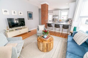 A modern living room with a TV, sofa, coffee table, and a kitchen in the background. - Edgewood: Close to Yale, Hospital & Park (New Haven)