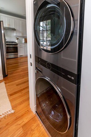 A compact washer and dryer unit with a stainless steel finish, located in a modern kitchen with white cabinets and a wooden floor.