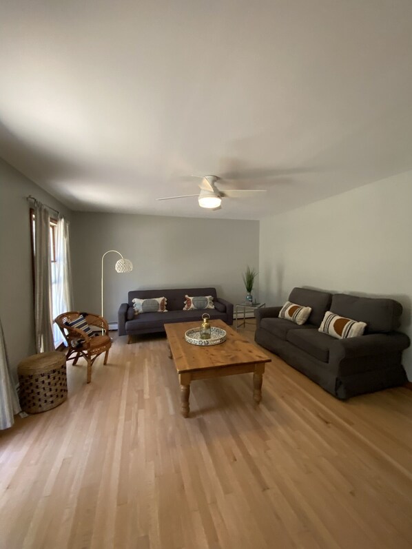 Una sala de estar con una mesa de centro de madera, dos sofás, un ventilador de techo y un ventanal con cortinas.