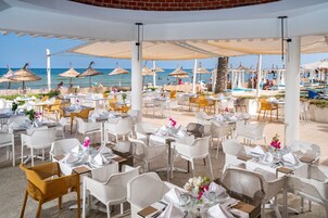 Un espacio exterior para comer con mesas, sillas, sombrillas y vistas a la playa y la piscina.