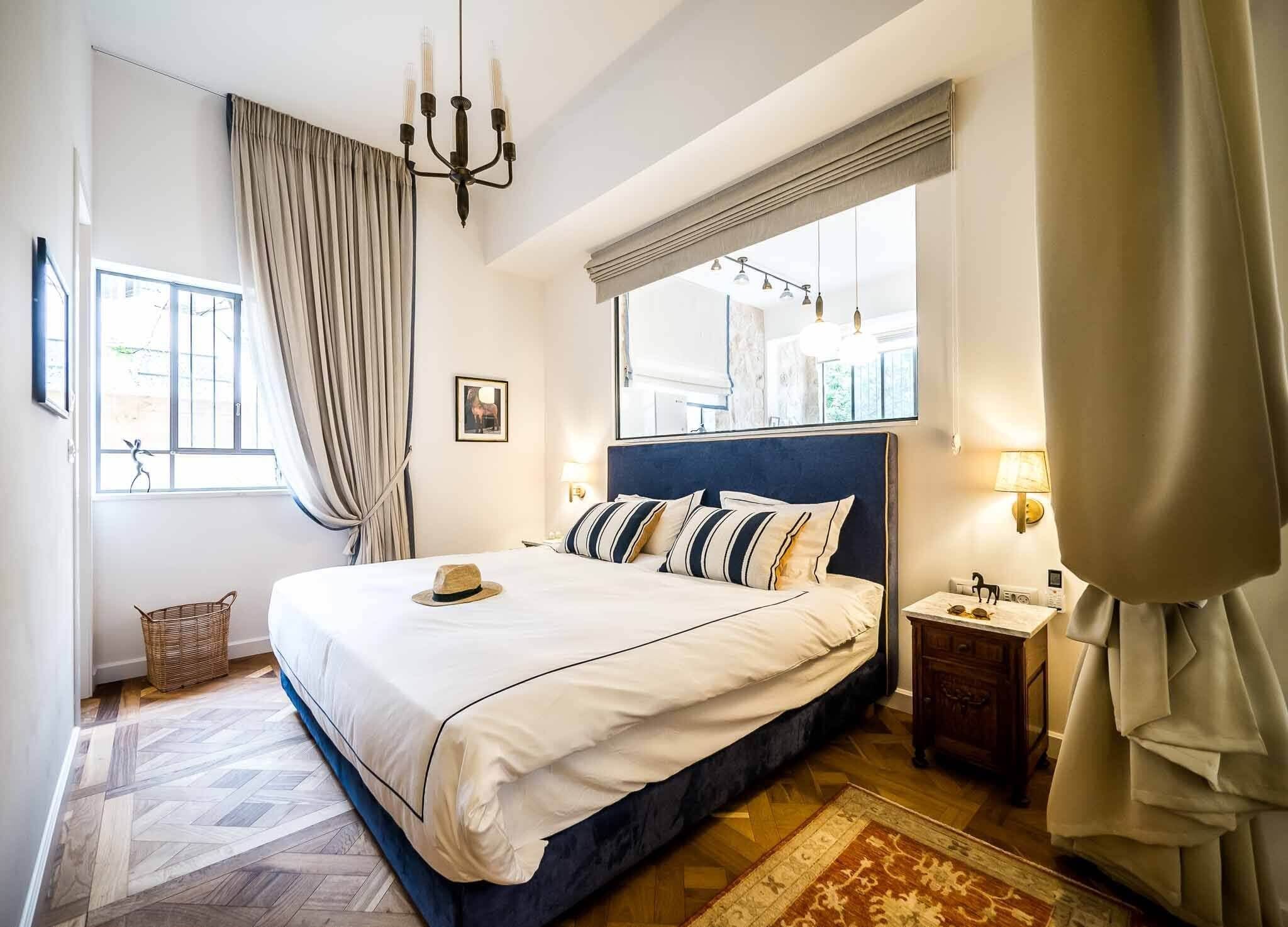 A hotel room with a large bed, a chandelier, a mirror, a nightstand, and a view of the outside.
