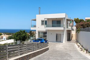 Une maison moderne à deux étages, avec une voiture garée dans l’allée, une vue dégagée sur la mer et un balcon doté d’une balustrade en verre.