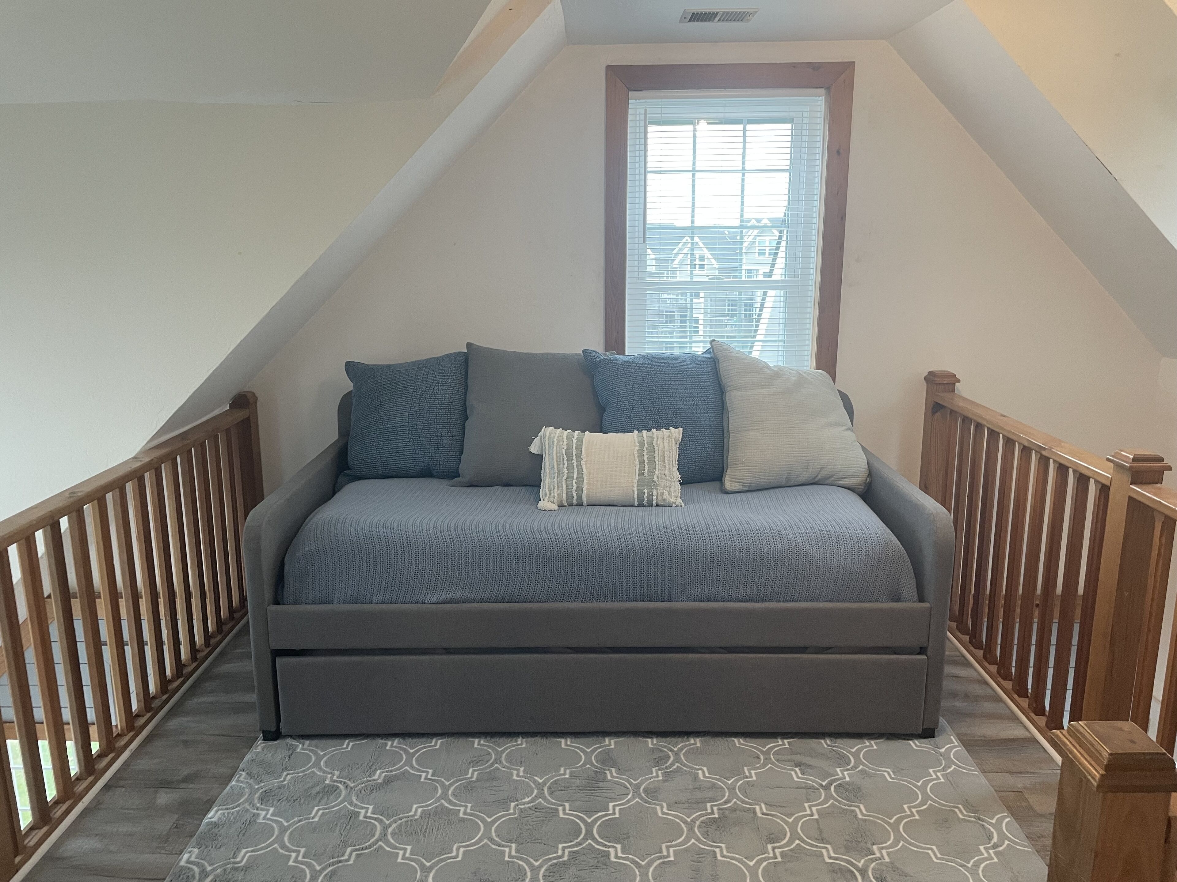 Ein Schlafzimmer im Loftsstil mit grauem Schlafsofa, einem gemusterten Teppich, Holzbalkonen und einem Fenster mit Jalousien.