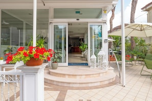 A hotel entrance with a glass door, a small outdoor seating area with chairs and tables, and potted plants. - Hotel Negresco (Riccione)