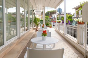 A covered outdoor seating area with white tables and chairs, potted plants, and a view of the street. - Hotel Negresco (Riccione)