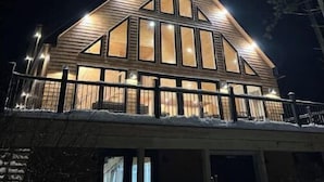 Een moderne, verlichte hut met een puntdak en grote ramen, omringd door bomen.