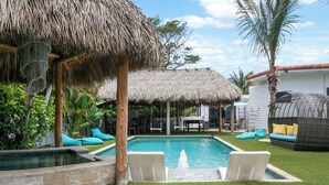 Una piscina con una estructura cubierta de paja, un área de descanso con cojines y un pequeño jardín con palmeras.