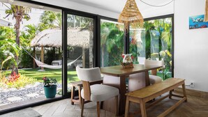 Comedor con mesa, sillas, banco, lámpara colgante y vista a un jardín con estructura de paja.