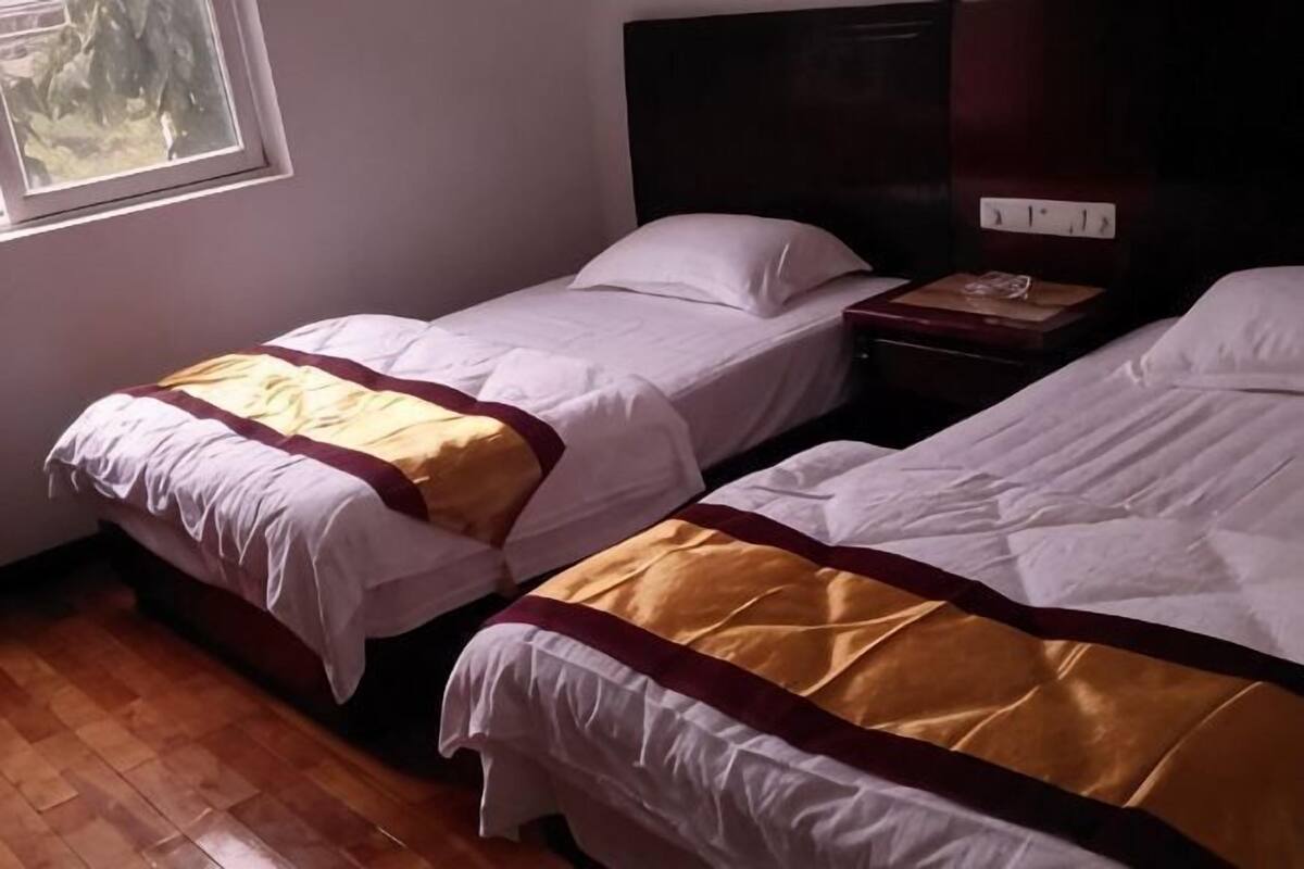 A room with two beds, wooden flooring, a window, and a nightstand with a lamp.