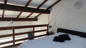 Un dormitorio con cama, mesita de noche, una pequeña mesa con lámpara y vistas a través de la barandilla de un balcón de madera.