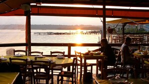 Un restaurante con mesas y sillas, vista al atardecer sobre el agua y un muelle a lo lejos.