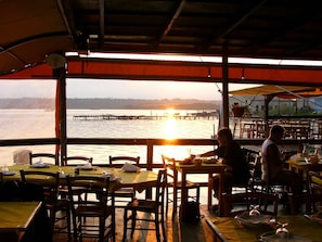 Un restaurante con mesas y sillas, vista al atardecer sobre el agua y un muelle a lo lejos.