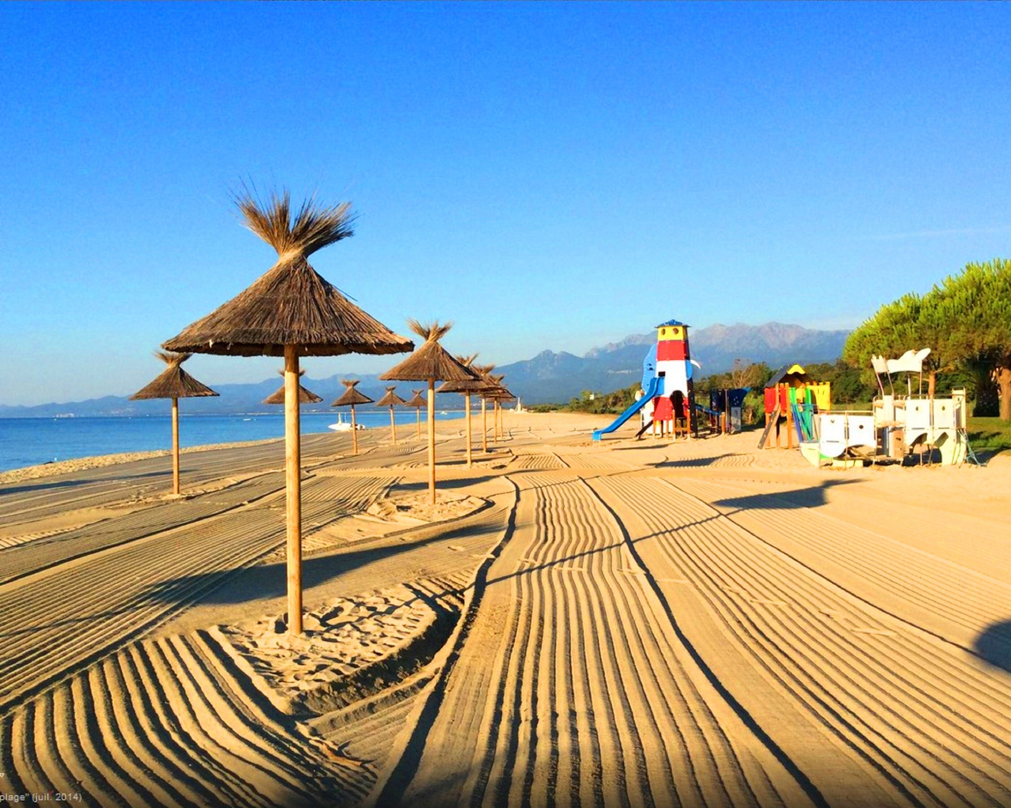 A beach with thatched umbrellas, a playground, and a clear view of the sea and mountains in the background.