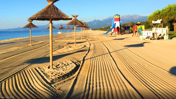 Una playa con sombrillas de paja, un área de juegos y una vista despejada del mar y las montañas al fondo.