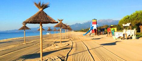 A beach with thatched umbrellas, a playground, and a clear view of the sea and mountains in the background.