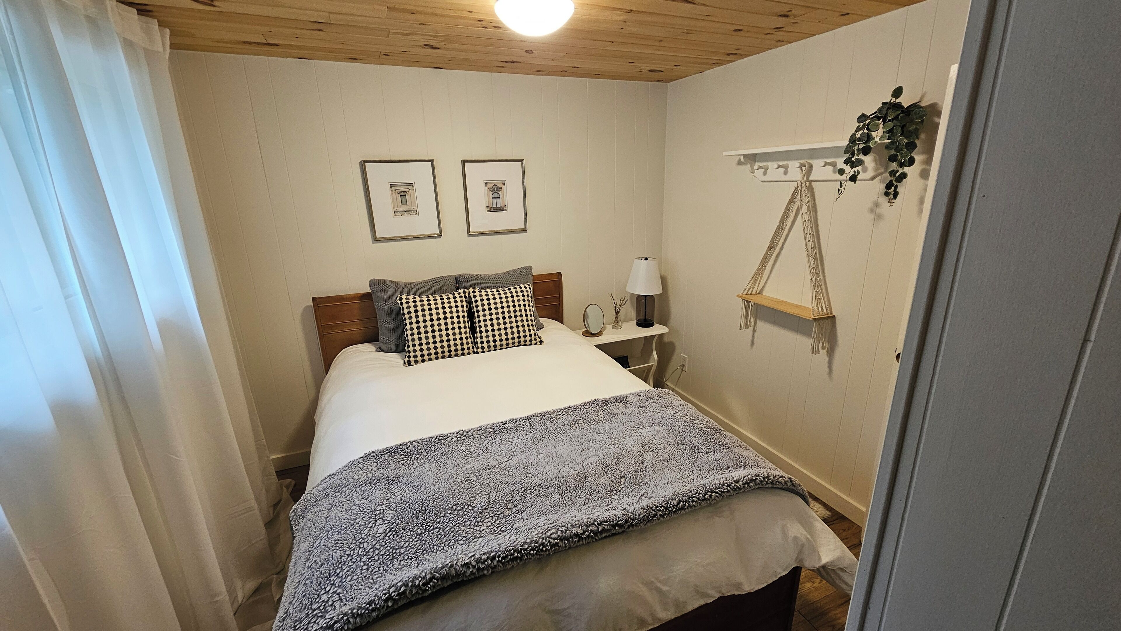 Une petite chambre à coucher accueillante, avec un plafond en bois, un lit agrémenté d’oreillers à motifs, une table de chevet avec une lampe et une plante fixée au mur.