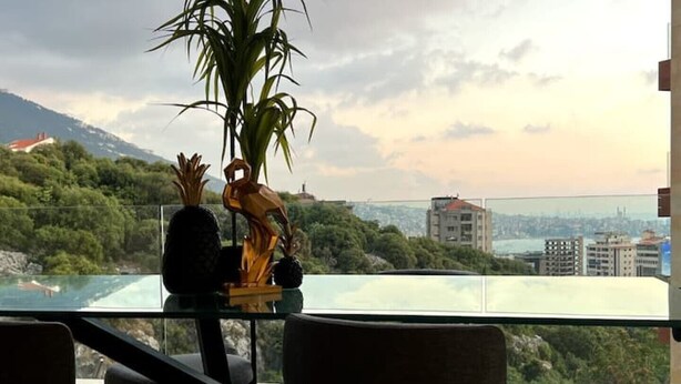 A modern room with a glass table, two chairs, a potted plant, and a view of a cityscape and mountains. - Modern Prestigious 24-hour Electricity 5 min from the Beach! (Ghazir)