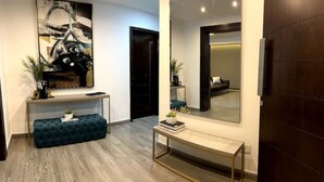 A modern hallway with a wooden floor, a dark blue bench, a console table with books and plants, and a large abstract painting on the wall. - Modern Prestigious 24-hour Electricity 5 min from the Beach! (Ghazir)