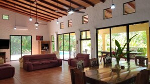 Un amplio espacio de sala con un sofá grande, una mesa de comedor y sillas, ventanas amplias que ofrecen vistas a zonas verdes y un ventilador de techo.