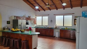 Una cocina con armarios de madera, un refrigerador, un microondas y un lavamanos. Hay taburetes en la barra y un ventilador de techo.