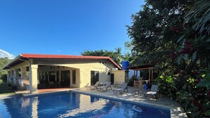 Zona de piscina con sillones, un porche cubierto y una casa con techo rojo.