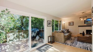 A spacious living room with a large sliding glass door leading to a wooden deck, a cozy sofa set, a fireplace, and a television mounted on the wall. - Hot Tub / Home Theater / Private Acre / Walk to Beach / Family and Dog friendly (Bainbridge Island)